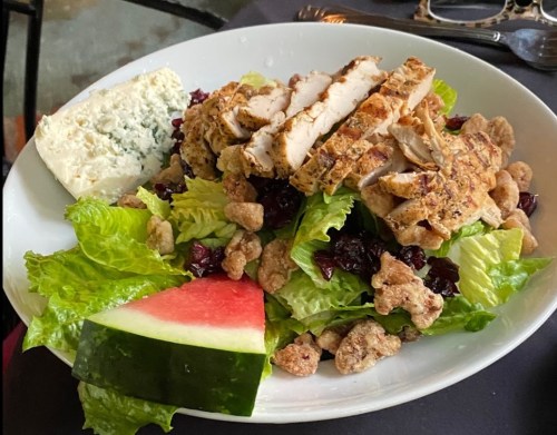 A plate of salad with sliced grilled chicken, lettuce, cranberries, candied nuts, and a wedge of watermelon.