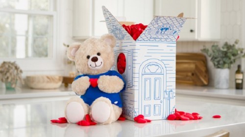A plush teddy bear in a blue shirt sits beside a decorative box filled with red rose petals on a kitchen counter.