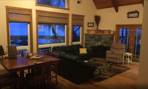 Cozy living room with a stone fireplace, large windows, and a dining table, illuminated by evening light.