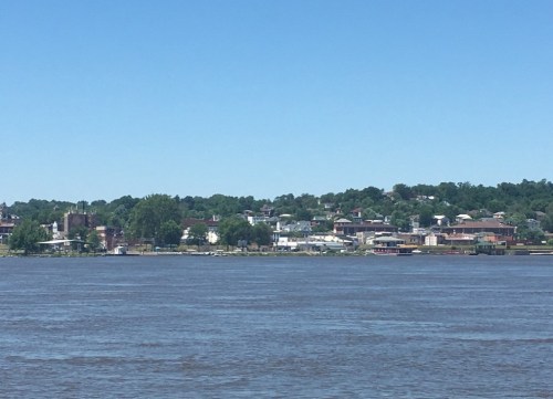 A view of a riverside town with houses and greenery under a clear blue sky. The river is calm and reflects the surroundings.