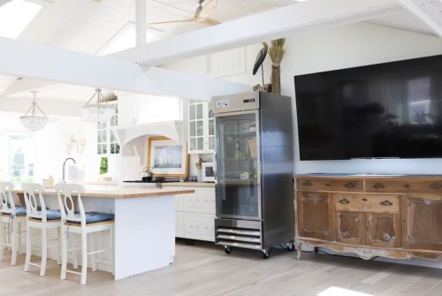 A bright kitchen featuring a stainless steel fridge, wooden dining table, and a large TV on the wall.
