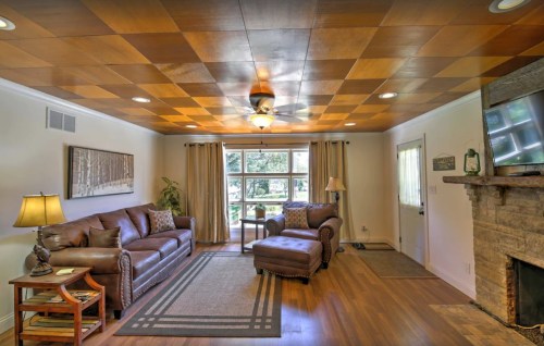 Cozy living room with a wooden ceiling, leather sofa, armchair, and a stone fireplace, featuring natural light from windows.
