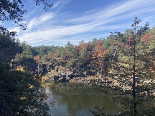 A serene river scene surrounded by colorful autumn trees and rocky cliffs under a blue sky with wispy clouds.