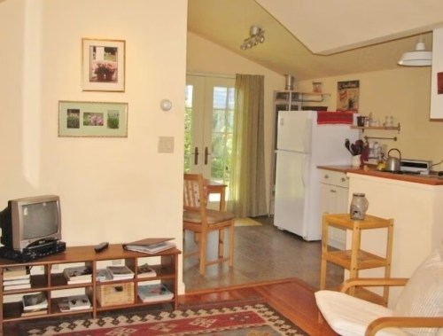 Cozy living space with a TV, bookshelf, and kitchen area featuring a white fridge and dining table. Natural light from windows.