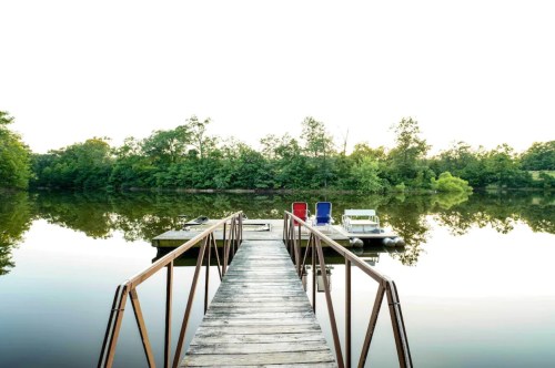 A wooden dock extends over a calm lake, surrounded by lush greenery and reflecting the serene sky.