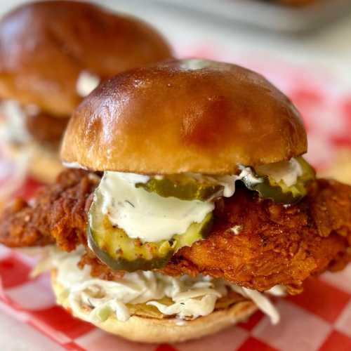 A close-up of a crispy fried chicken sandwich with pickles and creamy sauce on a soft bun, served with coleslaw.