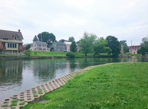 A serene river scene with grassy banks, houses lining the shore, and trees reflecting in the water under a cloudy sky.