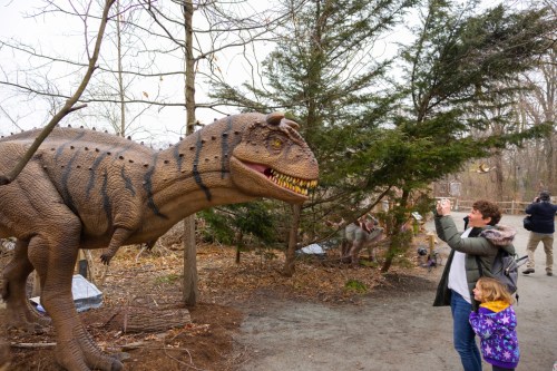 A woman and a child interact with a life-sized dinosaur model in a wooded area.