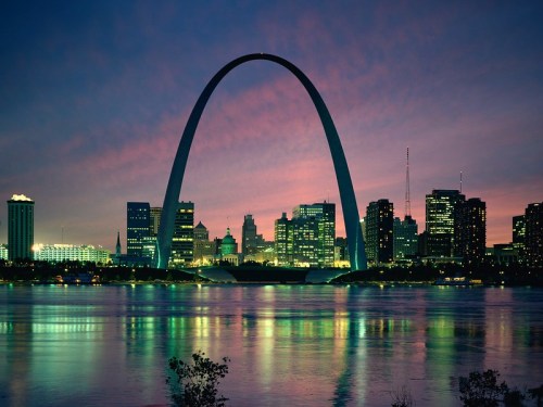 St. Louis skyline at dusk with the Gateway Arch reflecting in the water, surrounded by city lights and a colorful sky.