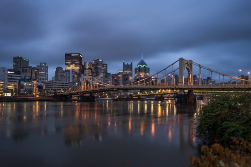 A city skyline at dusk, featuring a bridge over a river, with lights reflecting on the water and cloudy skies above.