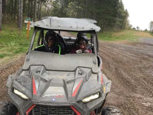 Two people wearing helmets smile while driving an off-road vehicle on a muddy trail surrounded by trees.