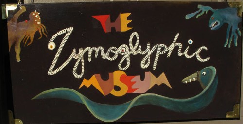 Colorful sign for "The Zymoglyphic Museum," featuring whimsical creatures and artistic lettering on a dark background.