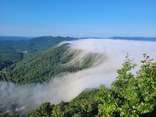 A panoramic view of lush green mountains shrouded in mist under a clear blue sky.