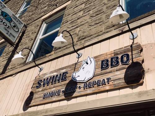 Sign for "Swine BBQ" featuring a pig illustration and the slogan "Smoke, Eat, Repeat" on a wooden background.