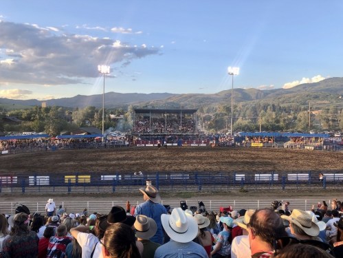A crowded rodeo arena with spectators wearing hats, surrounded by mountains and a clear blue sky.