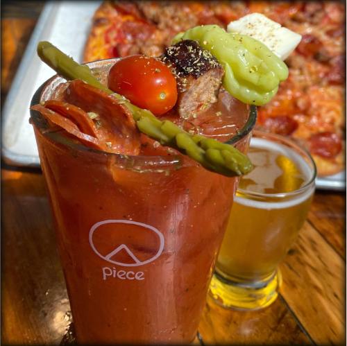 A vibrant Bloody Mary garnished with cherry tomatoes, pickles, and pepperoni, with a beer and pizza in the background.