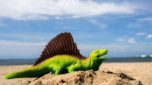 A green toy dinosaur with a spiky back sits on sandy beach, with a blue sky and water in the background.