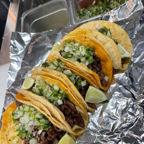 Five tacos filled with beef, topped with onions and cilantro, served with lime wedges on a foil-lined tray.