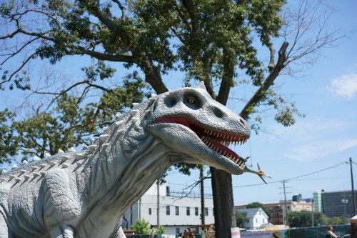A large, realistic dinosaur sculpture with sharp teeth, set against a blue sky and trees in the background.