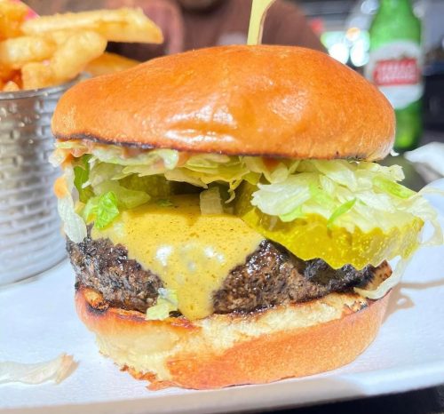 A close-up of a juicy cheeseburger with lettuce, pickles, and melted cheese on a toasted bun, served with fries.