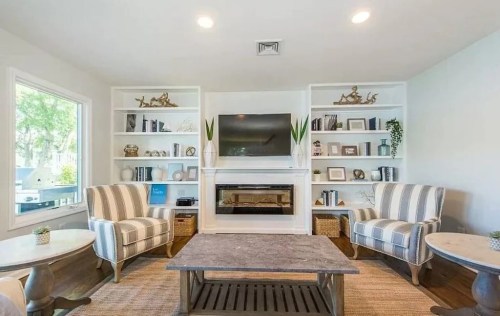 Cozy living room with striped chairs, a stone coffee table, and built-in shelves featuring decor and a TV above a fireplace.