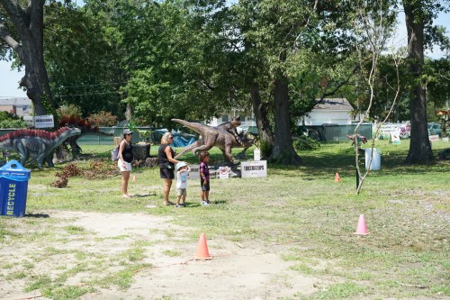Two children and an adult explore a dinosaur-themed outdoor exhibit with dinosaur sculptures and trees in the background.