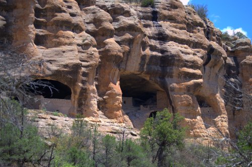 Ancient cliff dwellings carved into rocky cliffs, surrounded by trees and blue sky.