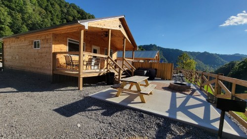 A wooden cabin with a porch, picnic table, and fire pit, surrounded by mountains and clear blue skies.