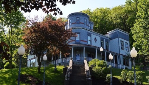 A large, historic house with a round tower, surrounded by trees and a grassy hill, featuring decorative architecture.
