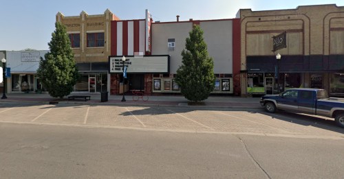 A street view of a small town with a movie theater, shops, and parked cars under a clear sky.