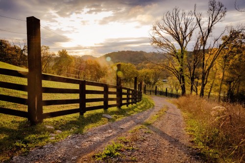 A winding gravel path leads through a scenic landscape with a wooden fence, trees, and a sunset in the background.