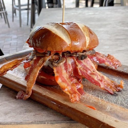 A pretzel bun burger overflowing with crispy bacon and toppings, served on a wooden board.