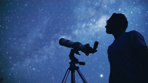 A silhouette of a person looking through a telescope at a starry night sky filled with stars and the Milky Way.