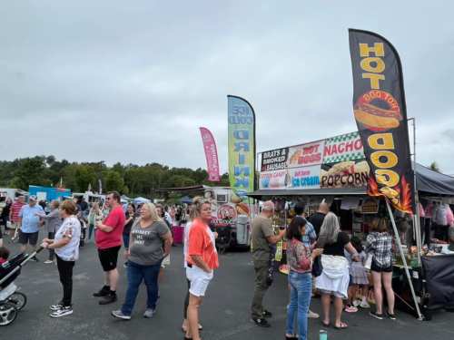 A busy food festival with people lined up at food stalls, colorful banners, and a cloudy sky in the background.