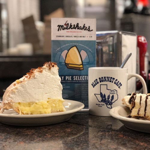 Two slices of pie with whipped cream on plates, alongside a menu and a coffee mug from Blue Bonnet Cafe.