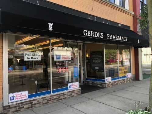 Exterior of Gerdes Pharmacy with a black awning, "OPEN" sign, and parking information visible on the windows.