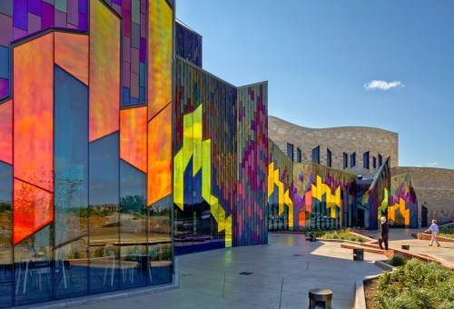 Colorful, modern building exterior with vibrant stained glass patterns and a clear blue sky. People walking nearby.
