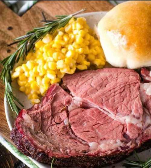 A plate with sliced roast beef, yellow corn, a soft roll, and a sprig of rosemary on a rustic wooden table.