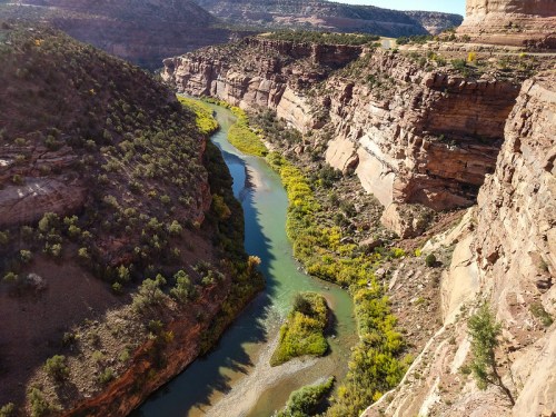 A winding river flows through a rocky canyon, surrounded by lush greenery and steep cliffs under a clear blue sky.