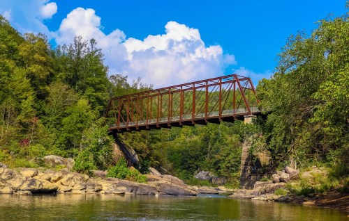 A rusted metal bridge spans a river, surrounded by lush greenery and rocky banks under a blue sky with clouds.