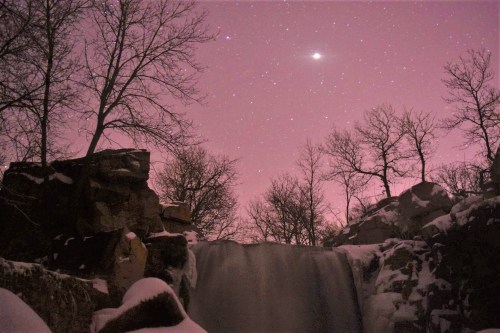 A serene waterfall surrounded by snow-covered rocks and trees, under a starry night sky with a hint of pink.
