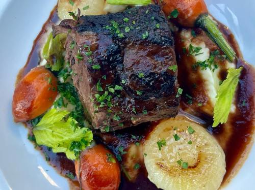 A plate of tender beef with roasted carrots, onions, and mashed potatoes, garnished with fresh herbs.