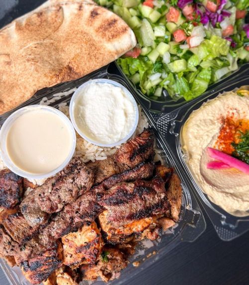 A takeout container with grilled meat, rice, hummus, tahini, pita bread, and a fresh salad on the side.