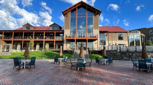A modern building with large windows, surrounded by outdoor seating and umbrellas, under a blue sky with clouds.