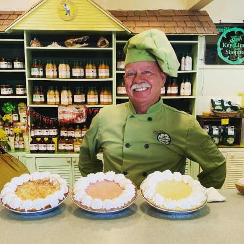 A smiling chef in a green uniform stands behind three pies on a counter, with shelves of products in the background.