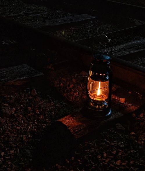 A glowing lantern sits on a wooden plank beside a railway track at night, casting a warm light on the surrounding stones.