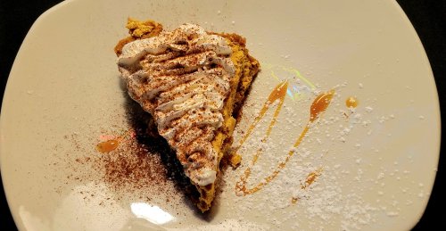 A slice of dessert topped with whipped cream, drizzled with caramel, and dusted with powdered sugar on a white plate.