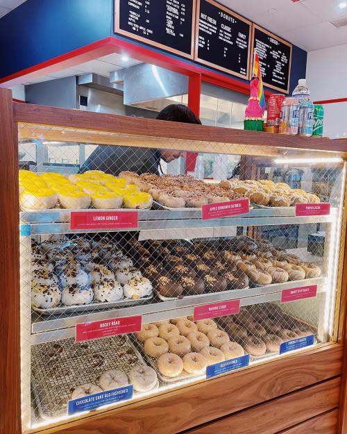 A display case filled with various donuts, including lemon ginger, apple cinnamon, and chocolate cake, in a bright shop.