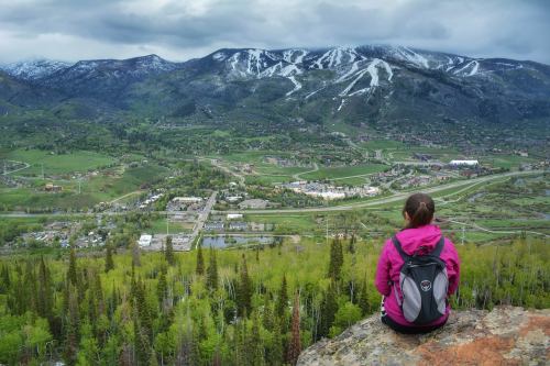 A person in a pink jacket sits on a rock, overlooking a lush valley and mountains with snow-capped peaks.