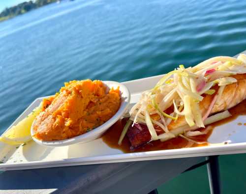 A plate with grilled fish topped with onions, served alongside sweet potato mash and a lemon wedge, with a lake in the background.